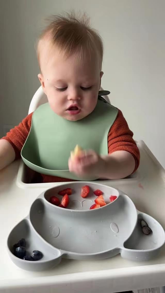 Baby in a high chair with a green bib and a bowl of fruits.